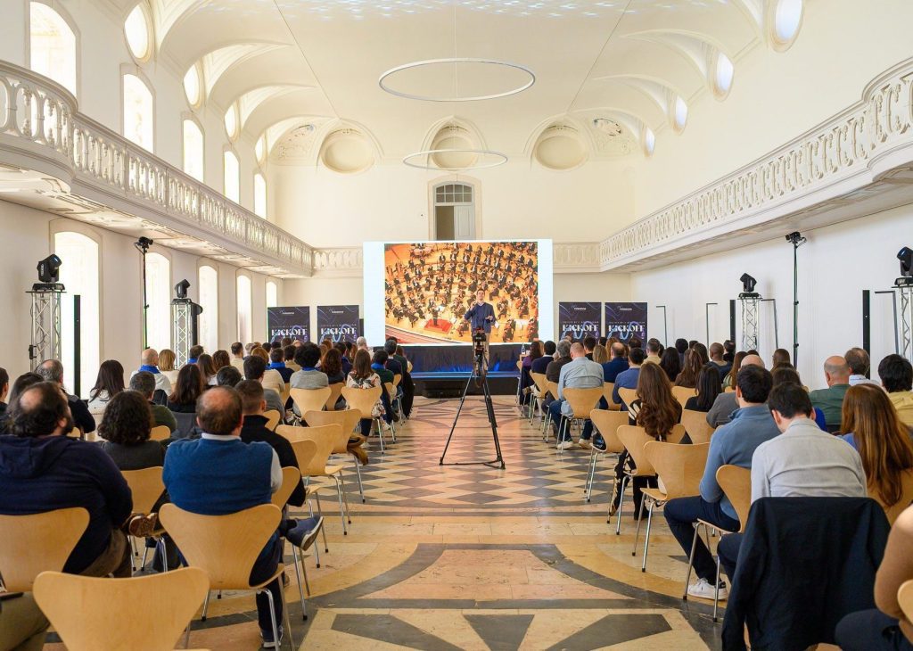 Conferência num salão histórico com plateia cheia e orador em palco diante de um ecrã