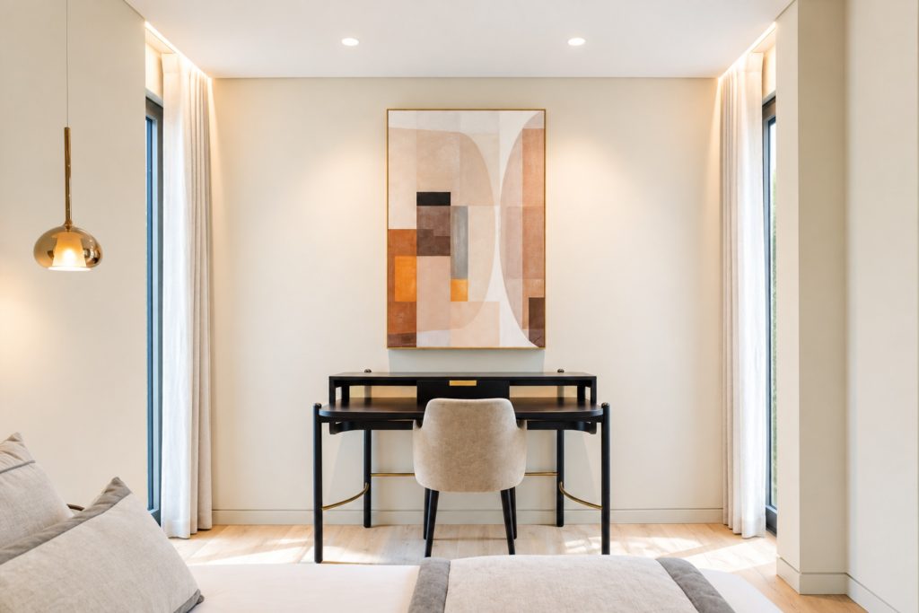 Quarto com secretária em madeira escura, cadeira estofada, quadro abstrato e cortinas de linho