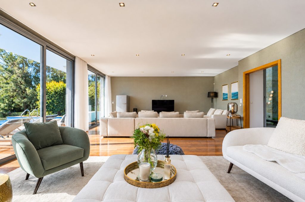 Sala de estar ampla com sofás claros, poltrona verde e janelas de vidro abertas para o jardim e a piscina