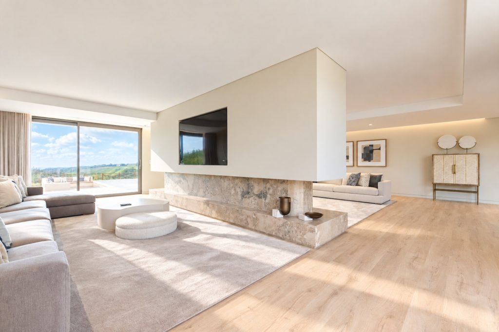 Sala ampla e luminosa com lareira central em mármore, sofás claros e janela com vista para o campo