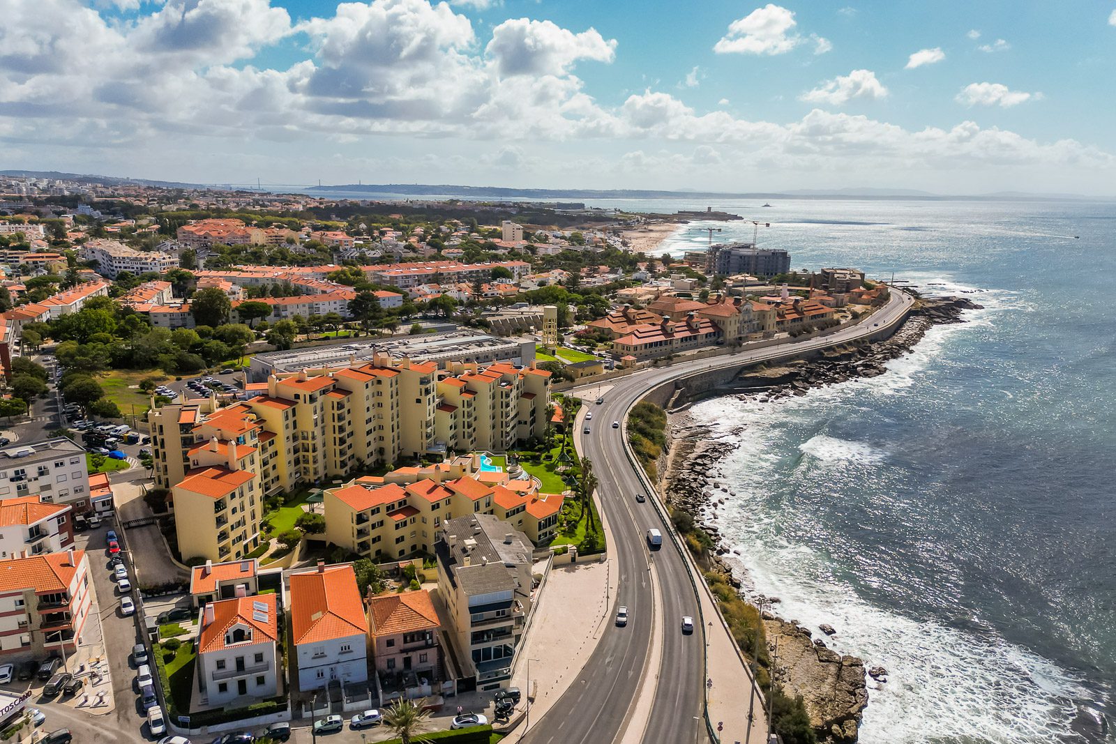 Vista aérea de drone sobre a costa, com condomínio com piscina, estrada marginal junto ao mar e a cidade em fundo